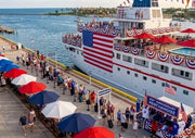 Large ship docked at a pier with people and umbrellas, featuring American flags and bunting.
